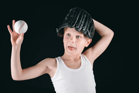 A child wearing a baseball glove prepares to throw a baseball, showcasing enthusiasm in a simple white tank top against a black background, illustrating the spirit of youth baseball and sparking curiosity about how much MLB ball boys girls make.
